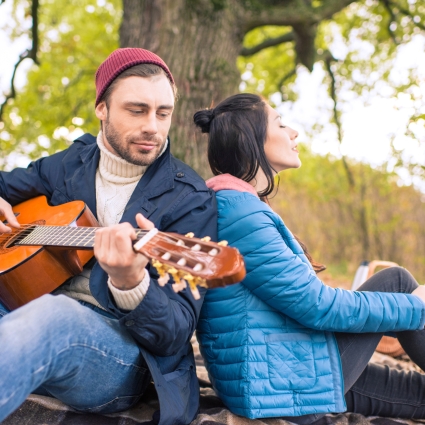 Happy romantic couple sitting on plaid and enjoying guitar in autumn forest