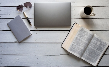 An overhead shot of an open bible near a laptop, coffee and a notebook with a pen on a white wooden surface