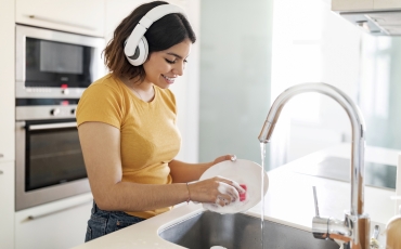 Young Arab Woman Wearing Wireless Headphones Washing Dishes In Kitchen, Smiling Middle Eastern Female Cleaning Plate And Listening Favorite Music, Enjoying Making Domestic Chores, Free Space